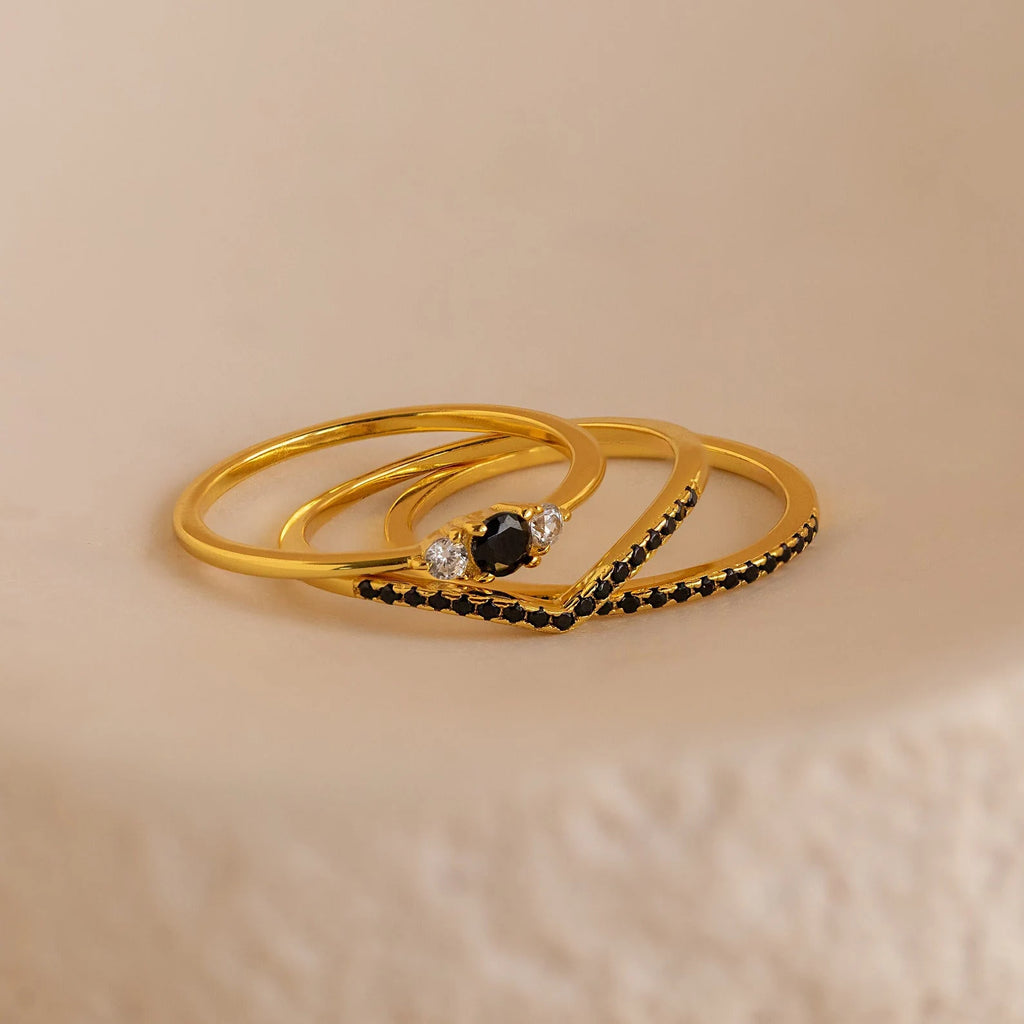 The Black Diamond Rings Set—three delicate gold rings featuring a striking black center stone and clear side stones—rests on a textured beige surface, perfect for effortless jewelry layering.