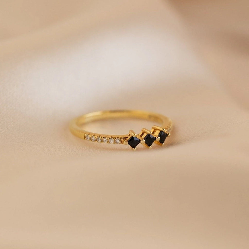 The Black Diamond Rings Set features a gold ring with three black stones and small clear stones, styled for stacking, shown on a soft beige fabric background.