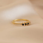The Black Diamond Rings Set features a gold ring with three black stones and small clear stones, styled for stacking, shown on a soft beige fabric background.