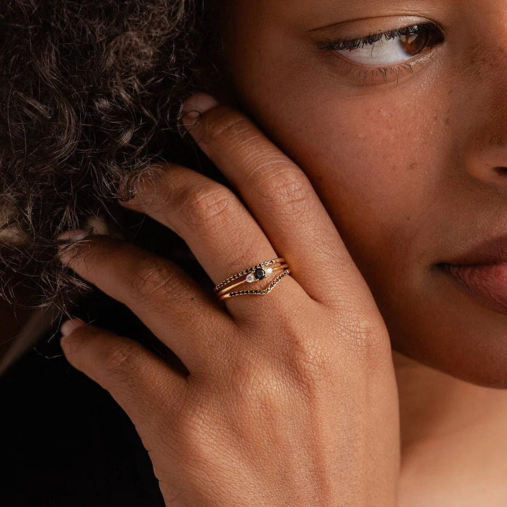A person with curly hair wears layered gold rings, including the Black Diamond Rings Set adorned with dark stones and delicate details, on their finger.