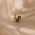 Two Onyx Flower Studs with small gold and black flower-shaped designs rest on a soft, cream-colored fabric background.