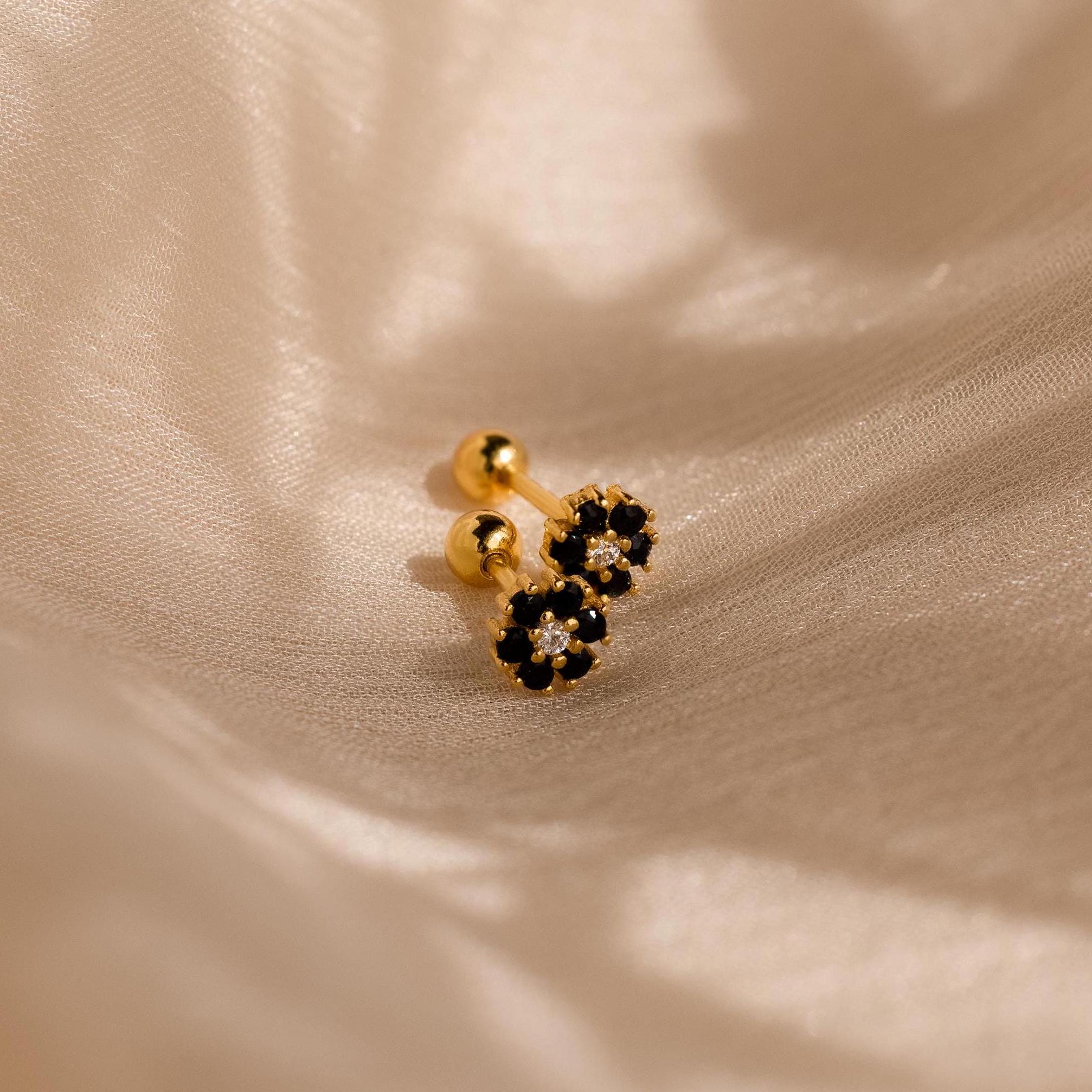 Two Onyx Flower Studs with small gold and black flower-shaped designs rest on a soft, cream-colored fabric background.