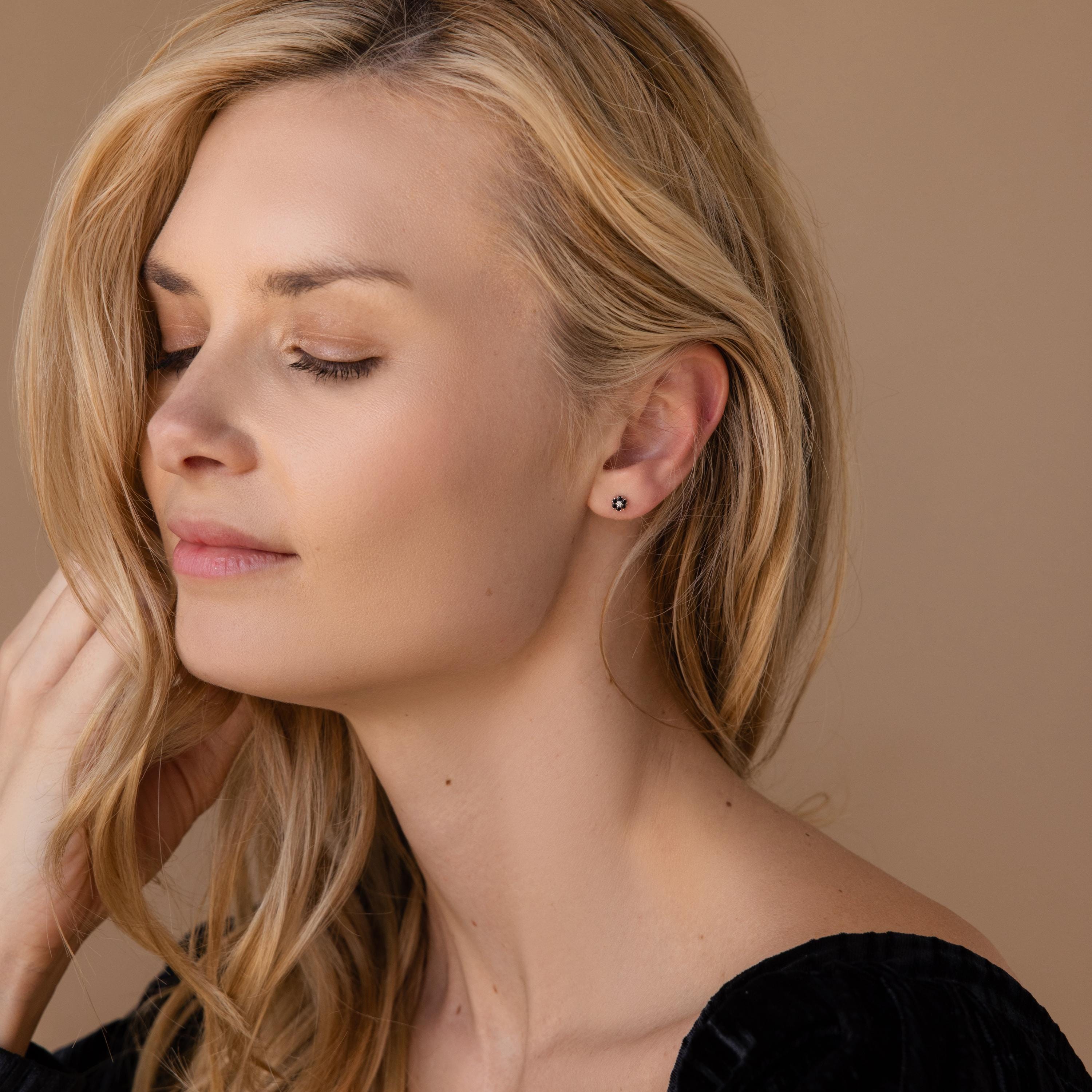 A woman with long blonde hair and closed eyes wears Onyx Flower Studs, elegantly posed against a beige background.