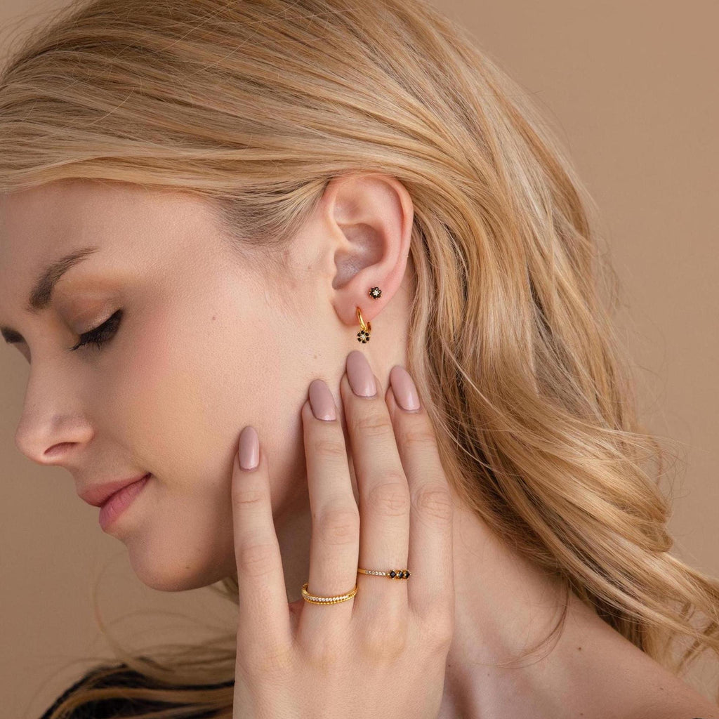 A blonde woman models the Onyx Flower Studs—gold and black onyx earrings—posing with her hand near her face against a beige background.
