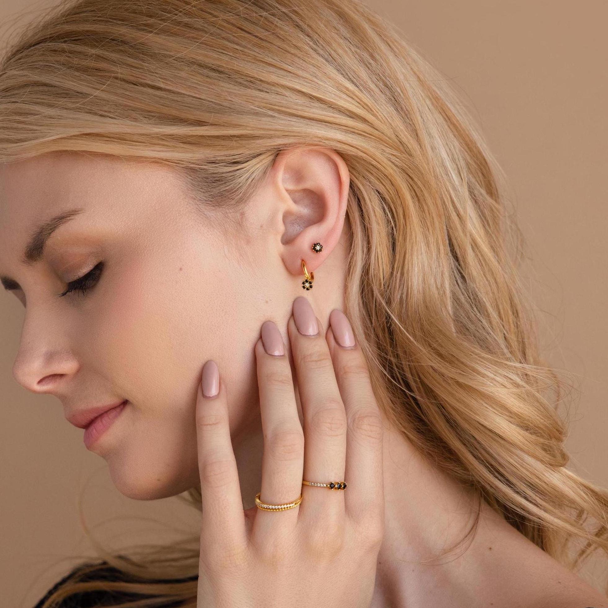A blonde woman models the Onyx Flower Studs—gold and black onyx earrings—posing with her hand near her face against a beige background.