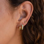 Close-up of an ear with gold hoops, sparkly gemstone earrings, a small gold stud, and Opal Bezel Huggies—delicate opal-adorned huggie earrings—set against wavy brown hair.