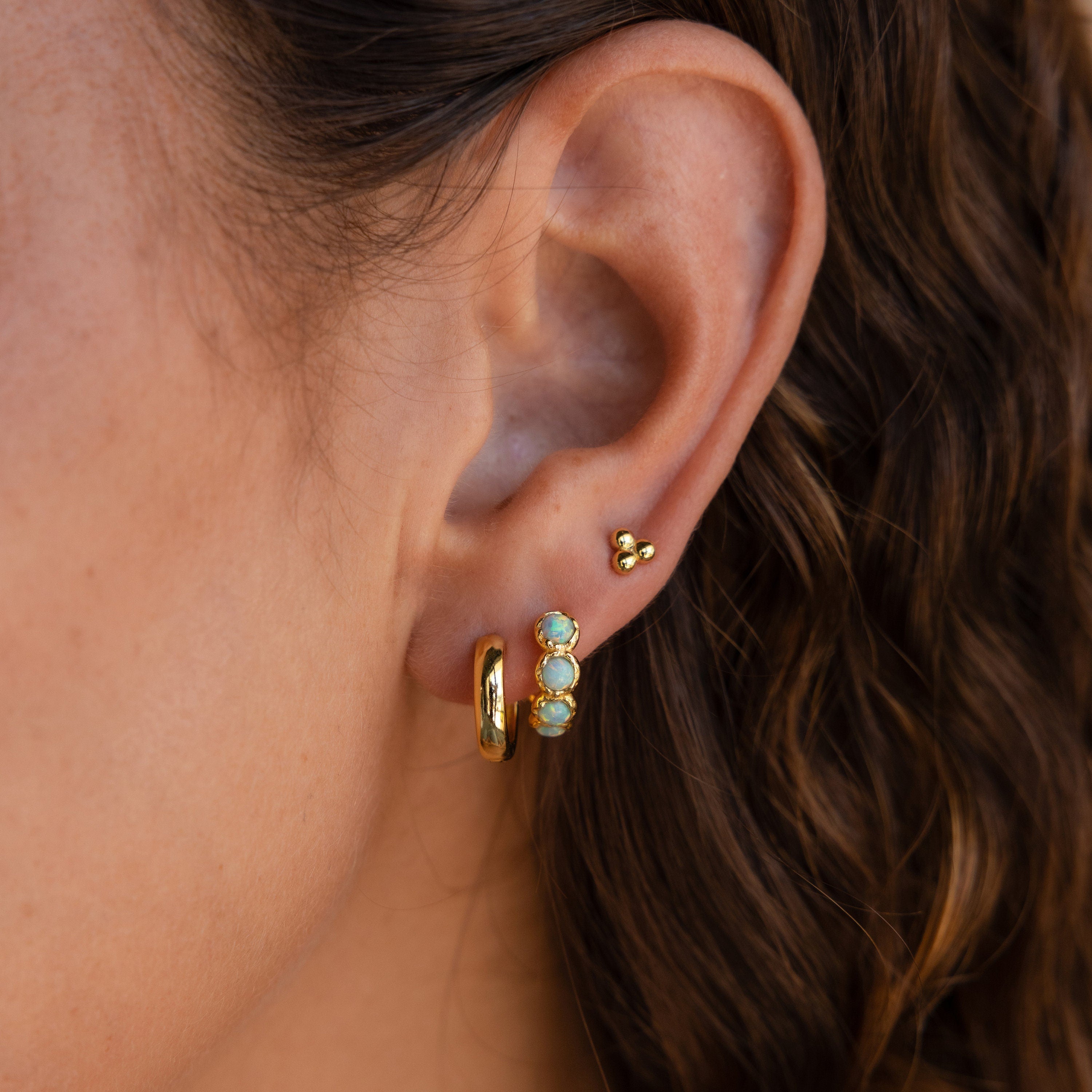 Close-up of an ear with gold hoops, sparkly gemstone earrings, a small gold stud, and Opal Bezel Huggies—delicate opal-adorned huggie earrings—set against wavy brown hair.