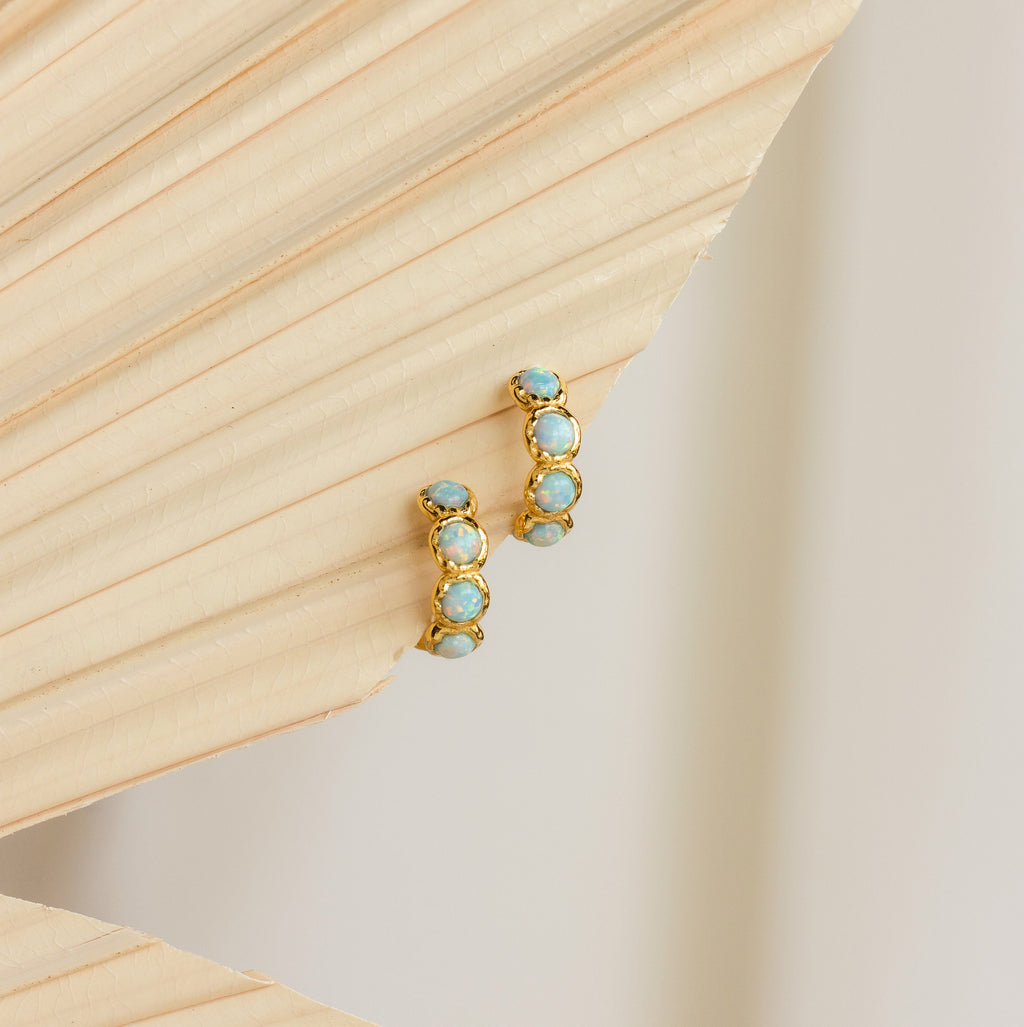 Opal Bezel Huggies earrings are displayed on a textured beige background.