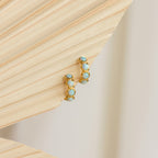 Opal Bezel Huggies earrings are displayed on a textured beige background.