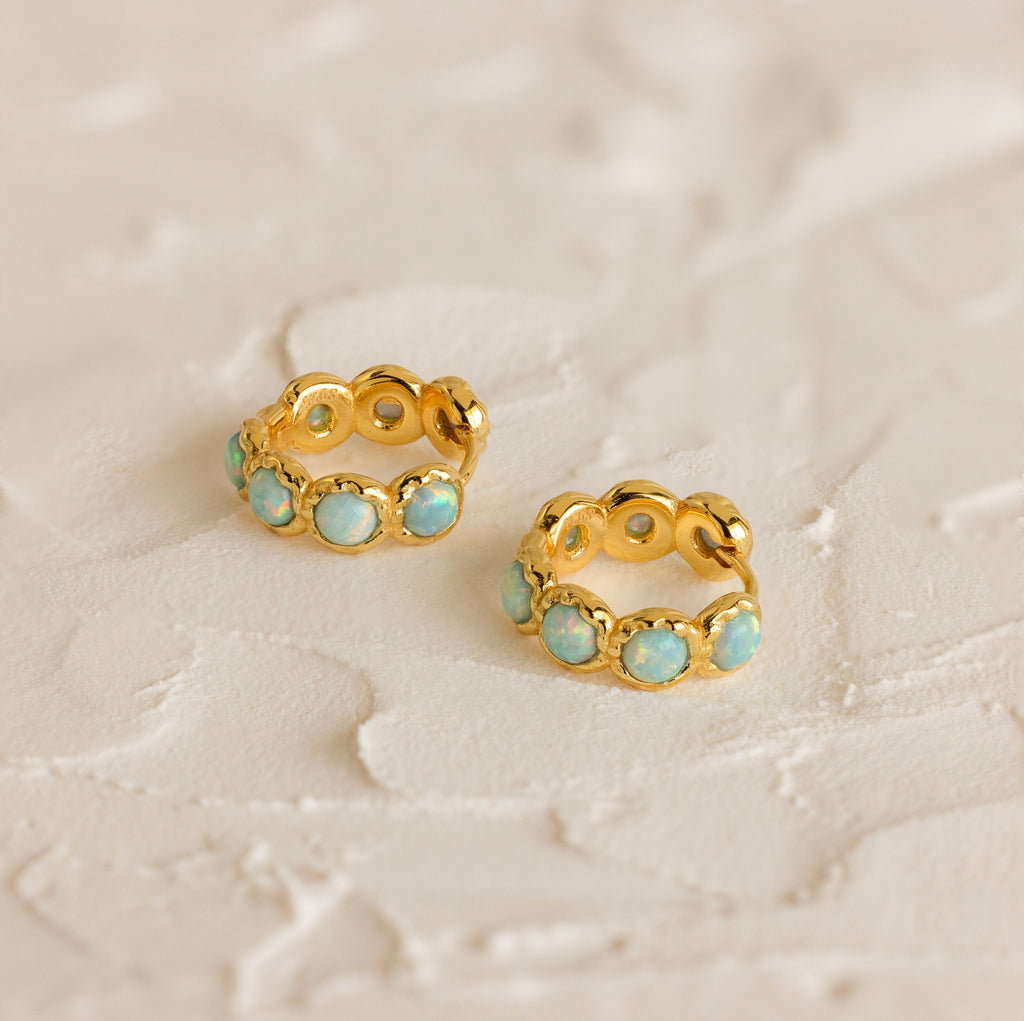 Two gold Opal Bezel Huggies featuring blue-green stones are displayed on a textured, off-white surface.
