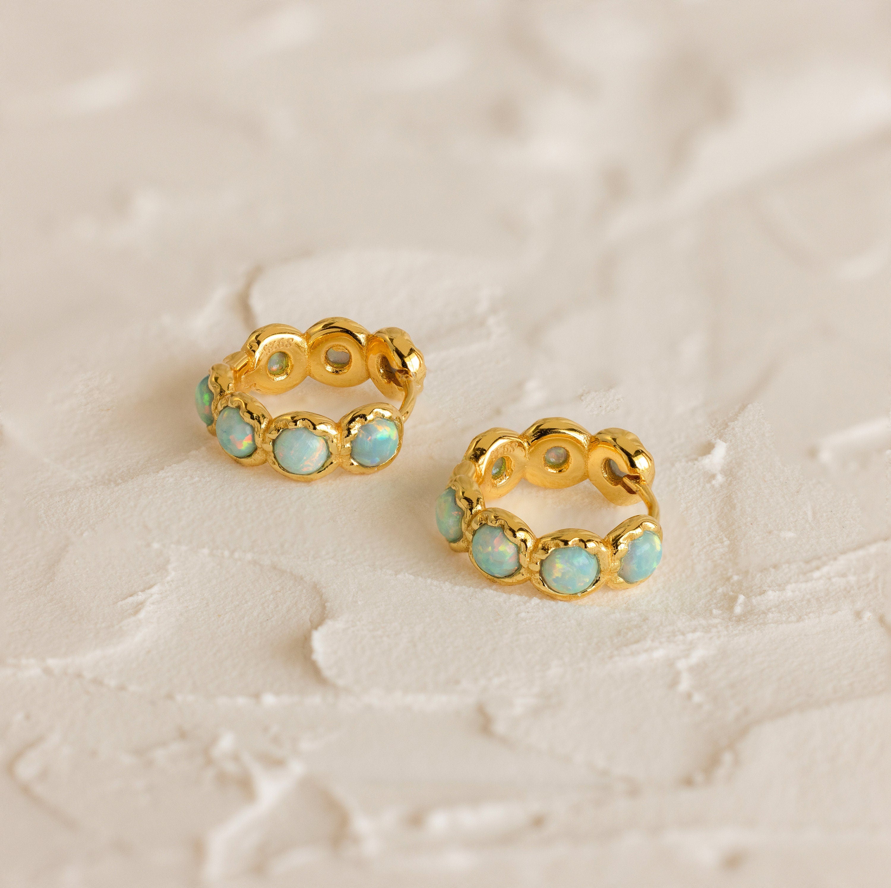 Two gold Opal Bezel Huggies featuring blue-green stones are displayed on a textured, off-white surface.