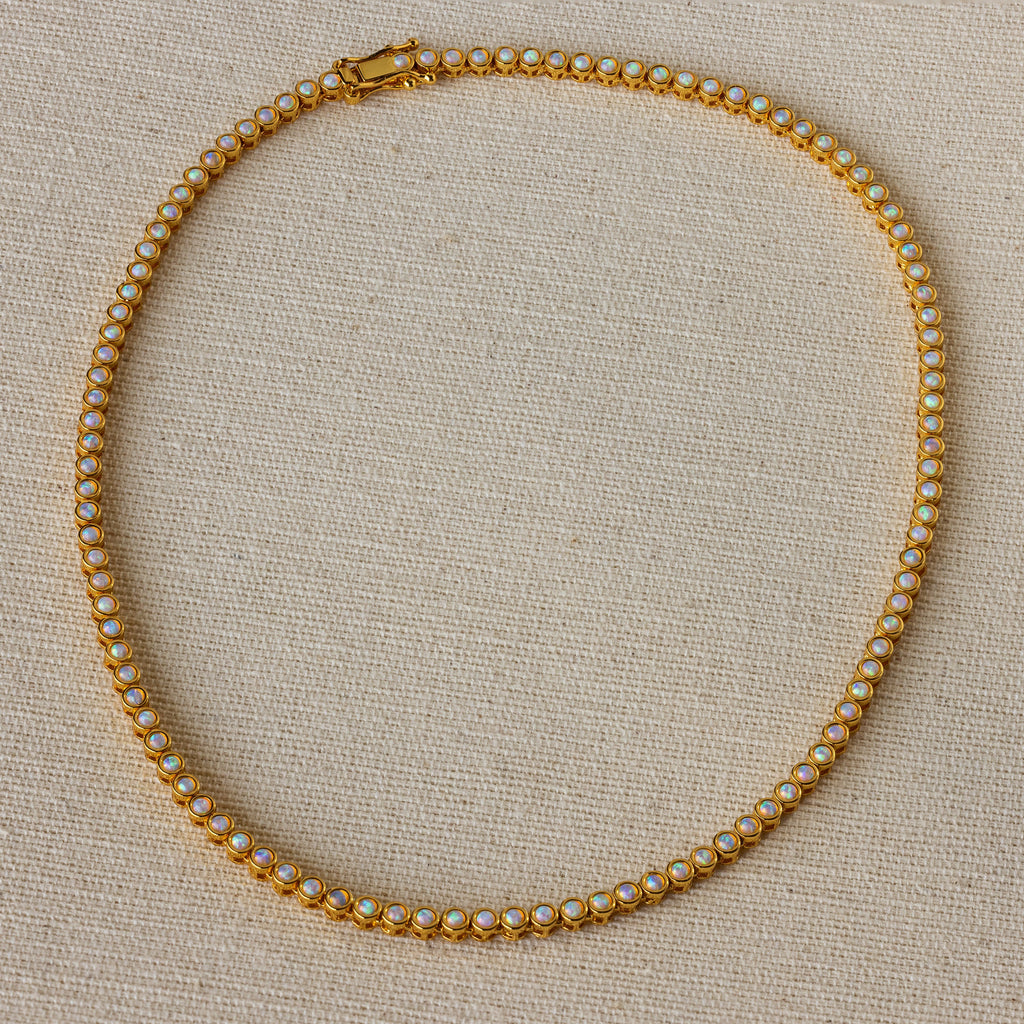 The Opal Bezel Tennis Necklace features small, round opals set in a single row, displayed on a beige textured surface.