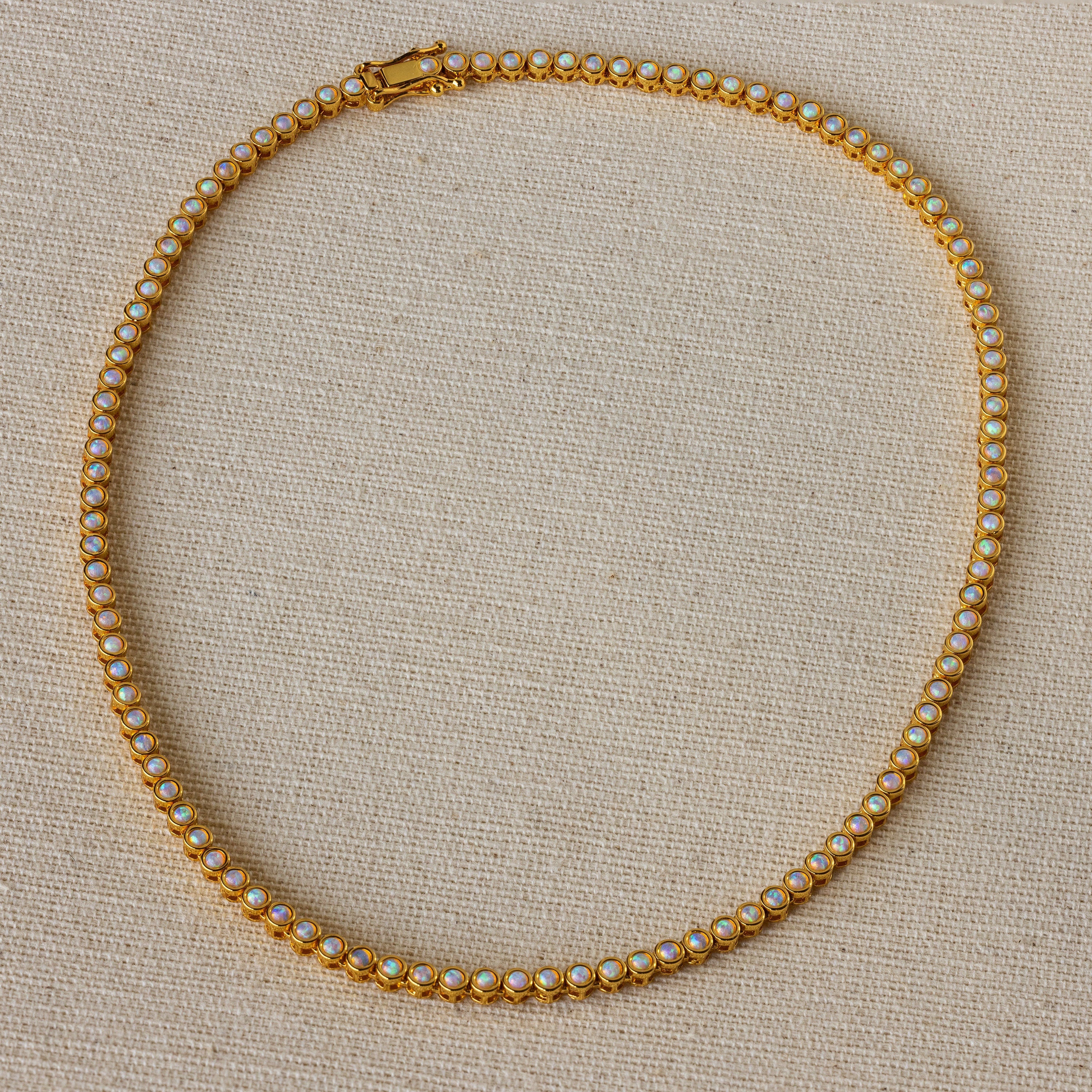 The Opal Bezel Tennis Necklace features small, round opals set in a single row, displayed on a beige textured surface.