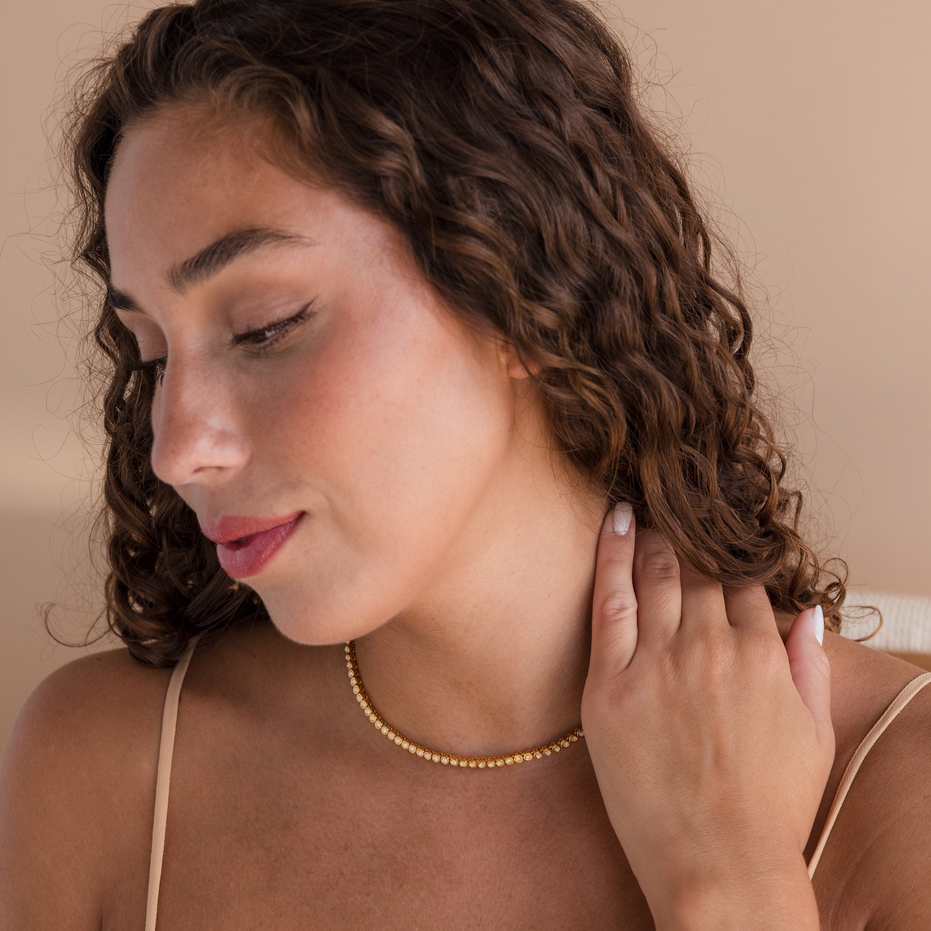 A woman with curly hair looks down, touching her neck while wearing the Opal Bezel Tennis Necklace in gold.
