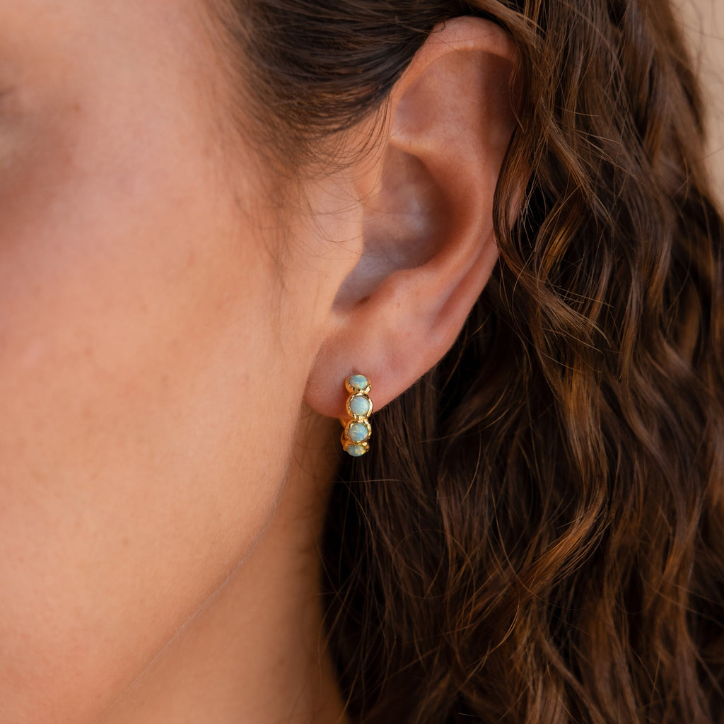 Close-up of a woman's ear wearing Opal Bezel Huggies, small gold earrings accented with turquoise stones.