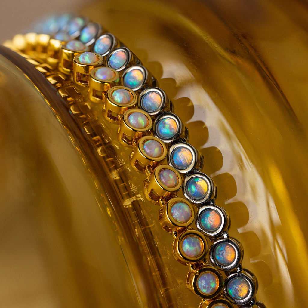The Opal Bezel Tennis Necklace, featuring iridescent opal stones set in gold, is displayed on a reflective golden-yellow surface, radiating timeless elegance.