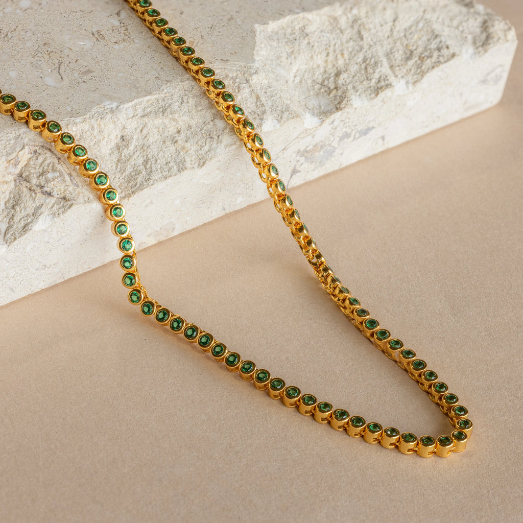 The Emerald Bezel Tennis Necklace features vibrant green gemstones, displayed on a beige surface with a light stone background, making this elegant piece a standout addition to any jewelry collection.