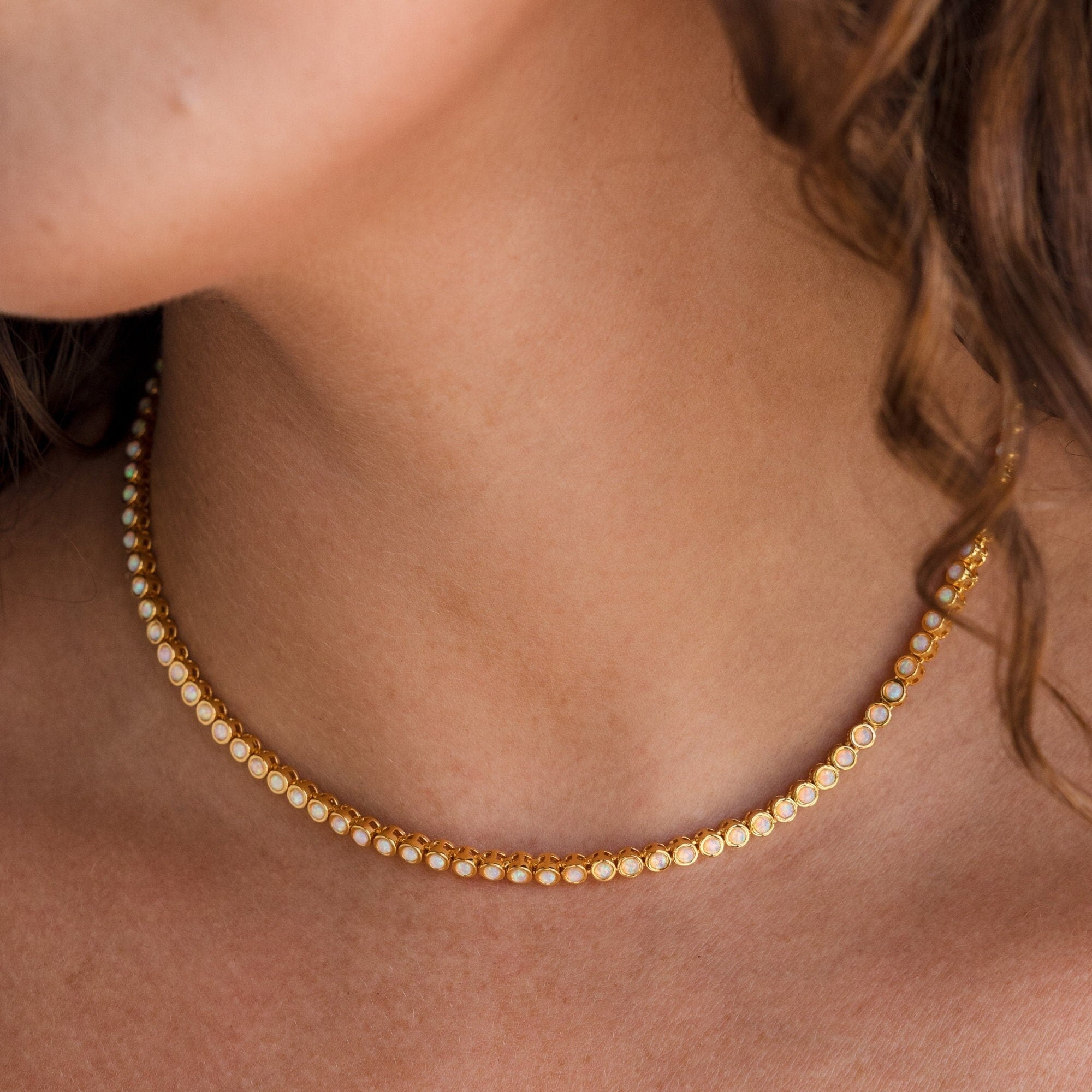 A close-up of a woman's neck adorned with the Opal Bezel Tennis Necklace, featuring small round opal gems set in gold.