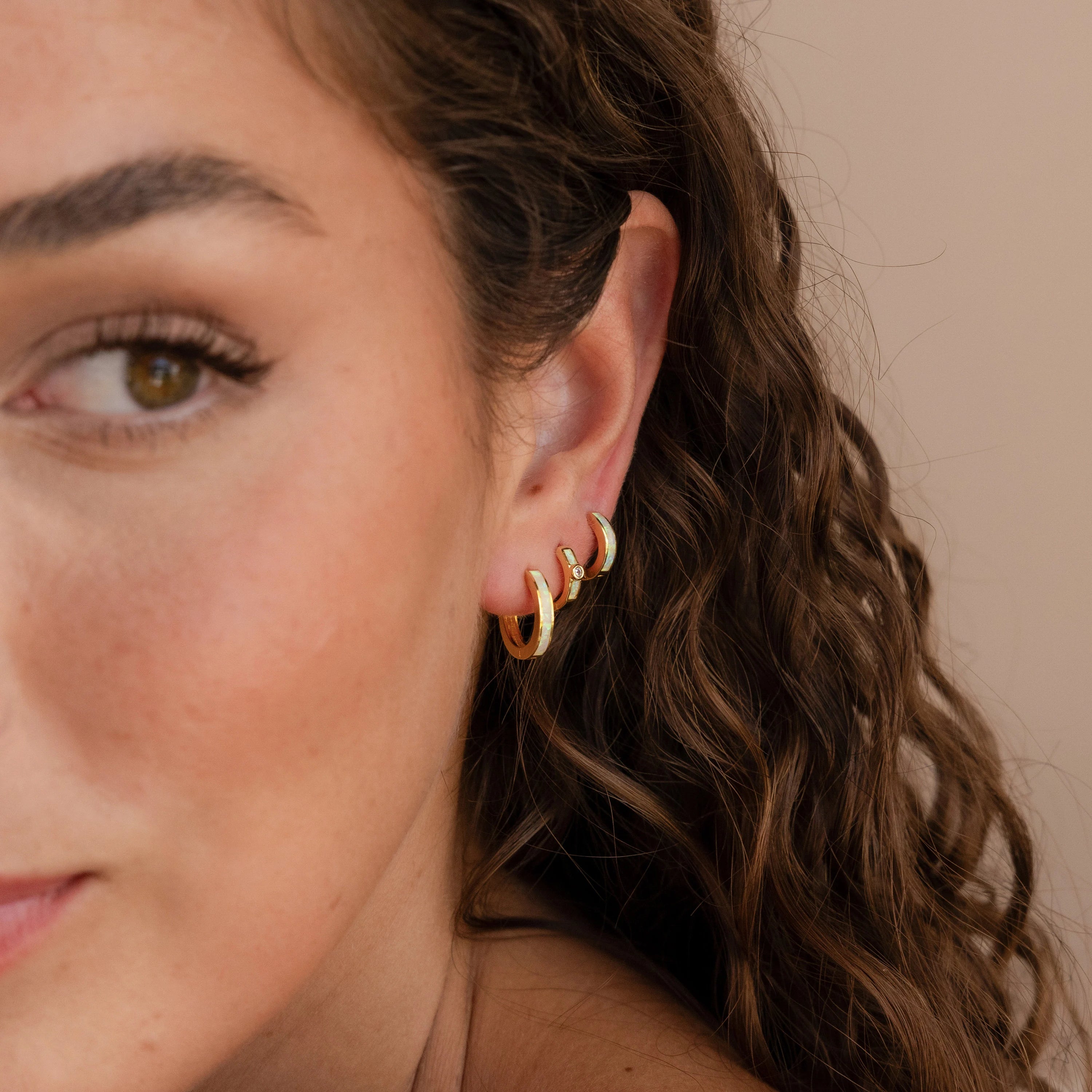 A woman with wavy brown hair wears three elegant gold hoop earrings, including the Opal Inlay Diamond Huggies, on her ear.
