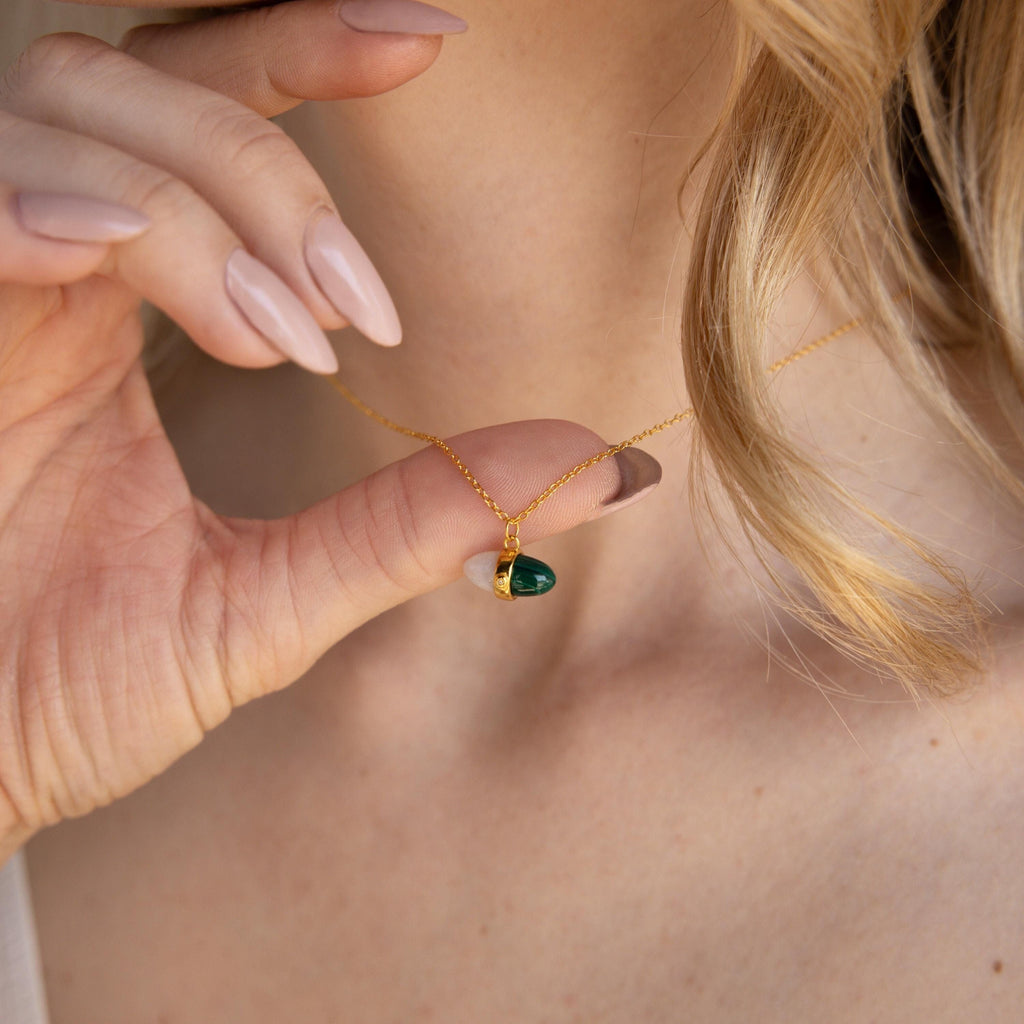 A woman holds the Malachite & Moonstone Necklace with a green teardrop pendant near her neck, displaying manicured nails and her love for healing gemstone jewelry.