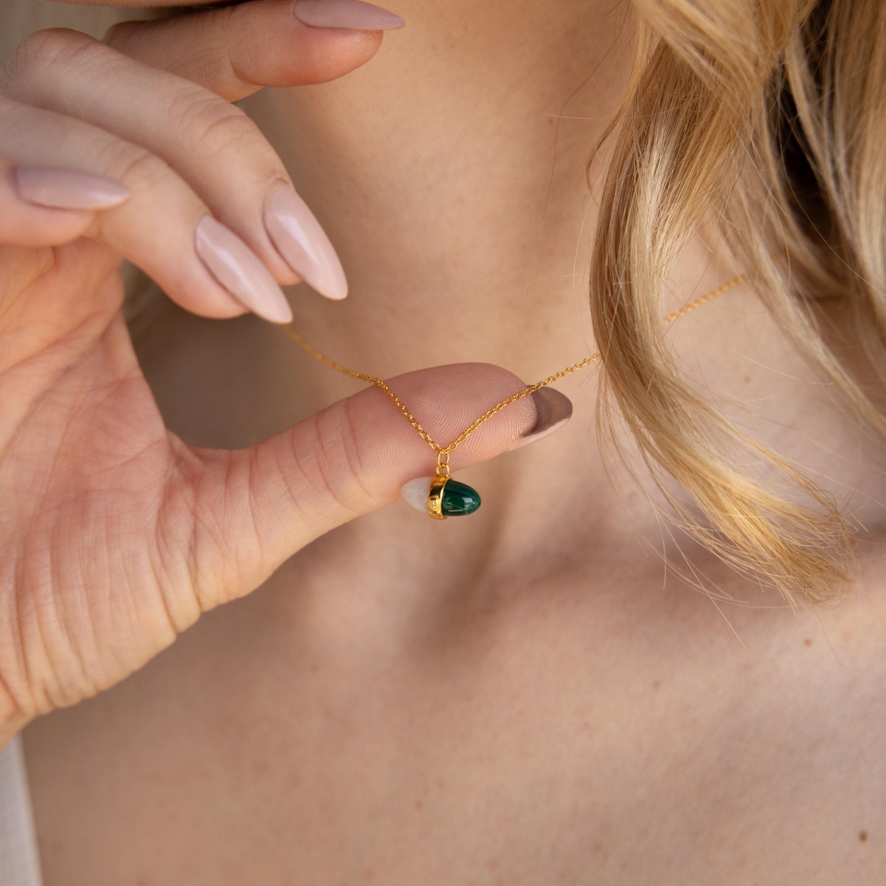 A woman holds the Malachite & Moonstone Necklace with a green teardrop pendant near her neck, displaying manicured nails and her love for healing gemstone jewelry.