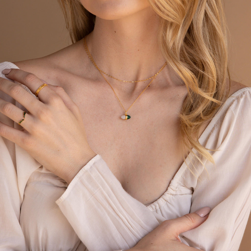 Woman in a cream blouse wears layered gold necklaces, including the Malachite & Moonstone Necklace, along with rings, as she touches her shoulder.