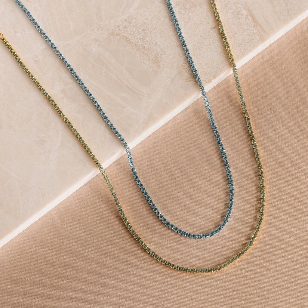 The Aquamarine Tennis Choker Necklace and a gold chain are displayed on a beige and marble surface—ideal as elegant wedding jewelry or a stunning bridal necklace.