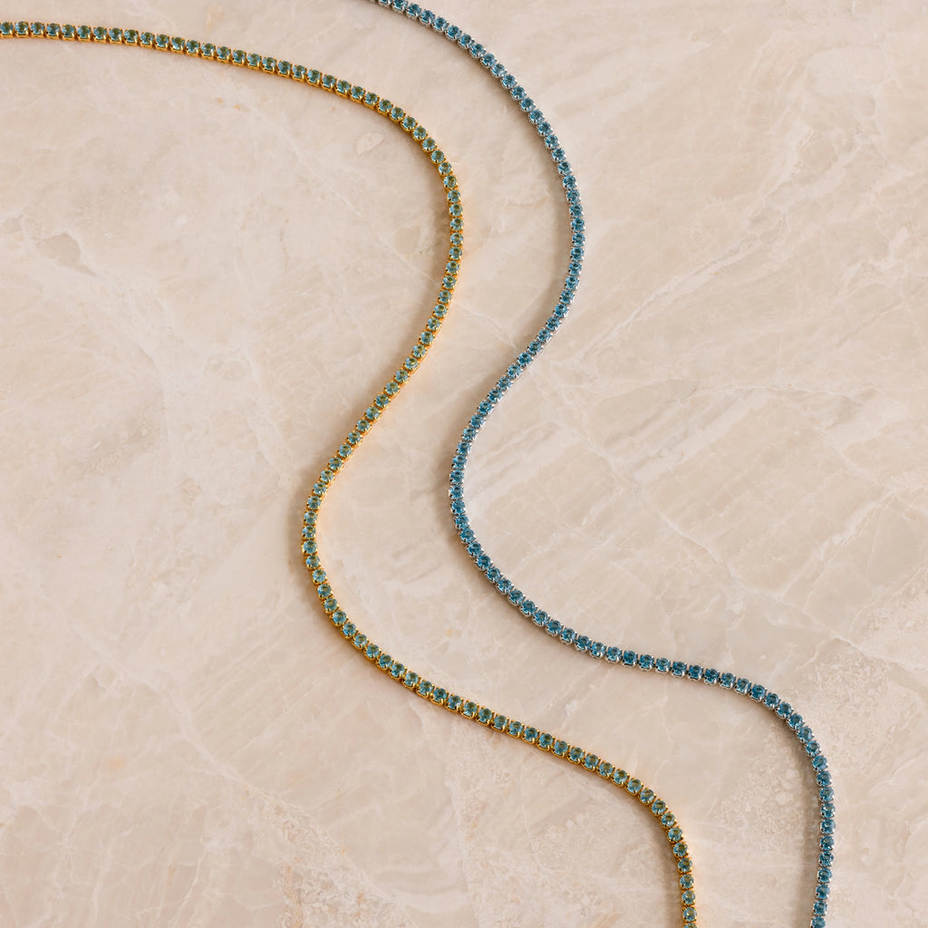 Two shimmering tennis necklaces—one gold and one Aquamarine Tennis Choker Necklace—are arranged in wavy lines on marble, perfect as wedding jewelry or a stunning bridal accessory.