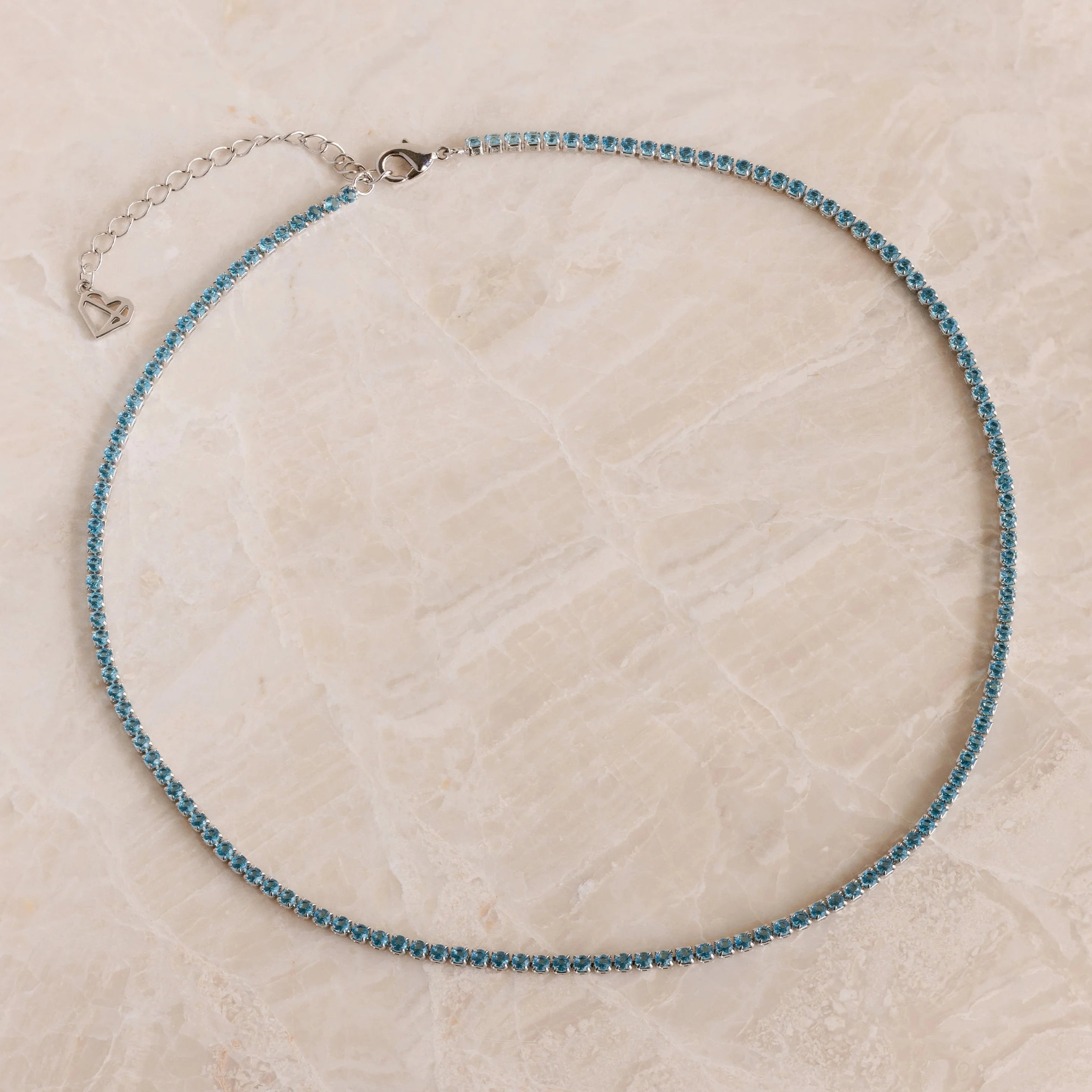 The Aquamarine Tennis Choker Necklace features a delicate silver chain with small blue stones and a heart charm extender, making it ideal as bridal or wedding jewelry. Shown elegantly on a marble surface.