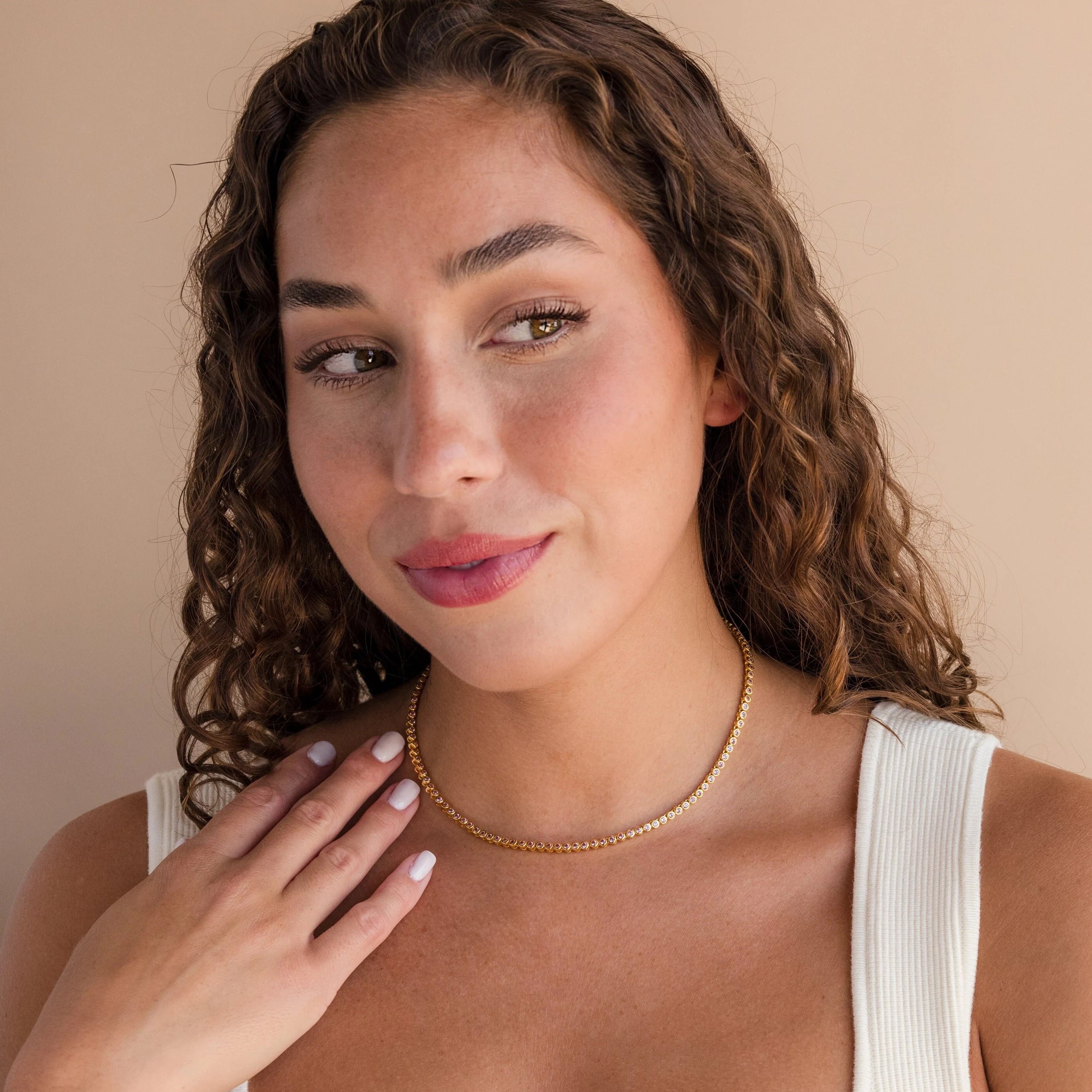 Woman with curly hair wears the Pink Bezel Tennis Necklace and a cream tank top, smiling softly against a beige background.