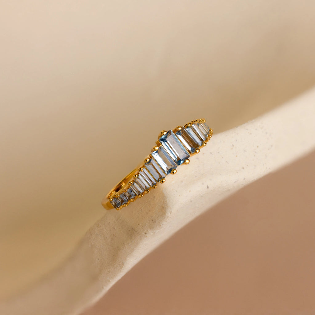 The Art Deco Aquamarine Ring, featuring a gold band and rectangular white gemstones, is displayed on a soft beige curved surface, radiating vintage glamour.