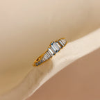 The Art Deco Aquamarine Ring, featuring a gold band and rectangular white gemstones, is displayed on a soft beige curved surface, radiating vintage glamour.