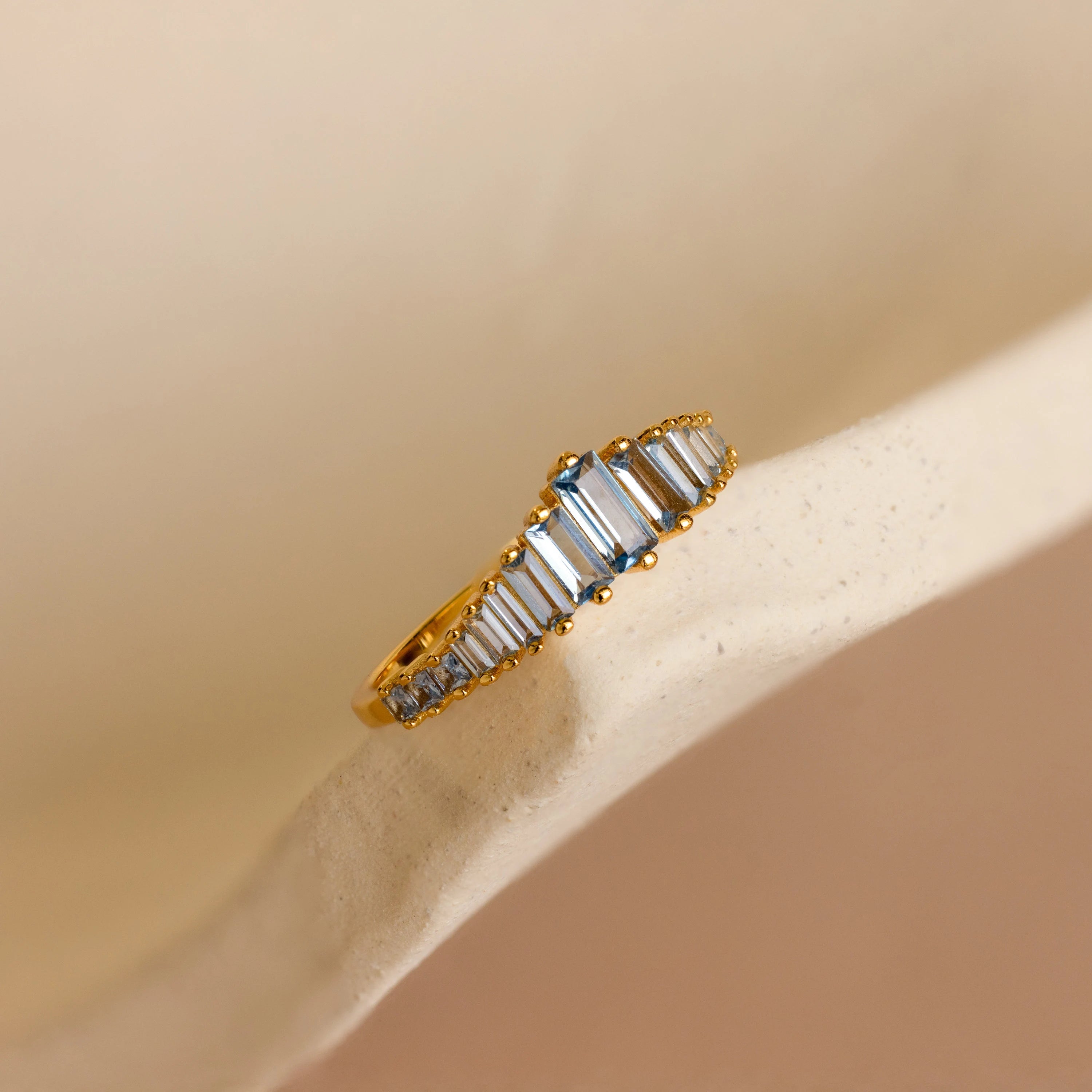 The Art Deco Aquamarine Ring, featuring a gold band and rectangular white gemstones, is displayed on a soft beige curved surface, radiating vintage glamour.