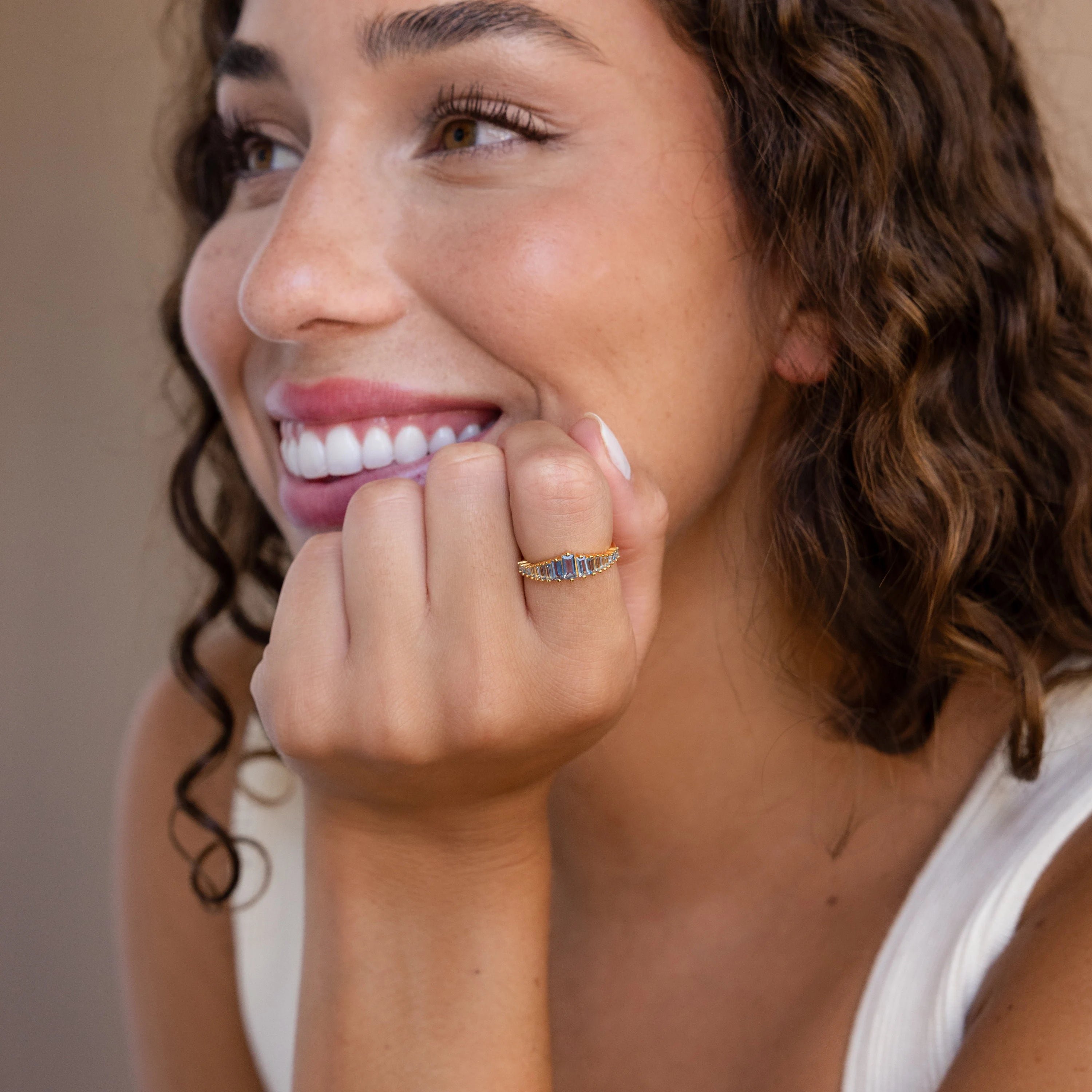 A smiling woman with curly hair rests her chin on her hand, showcasing the Art Deco Aquamarine Ring and its vintage gold sparkle.