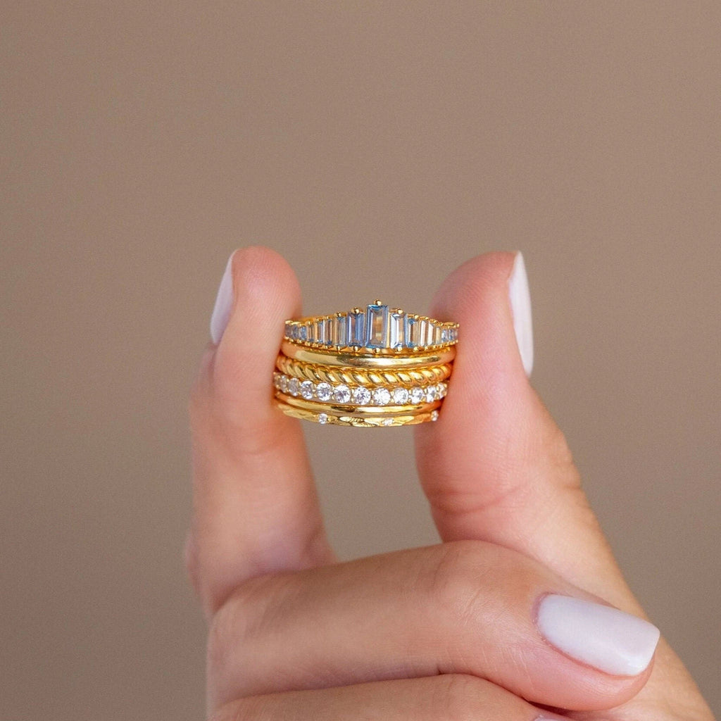 A hand with pale nails holds several stacked gold and diamond rings, including the Art Deco Aquamarine Ring, against a beige background for a hint of vintage glamour.