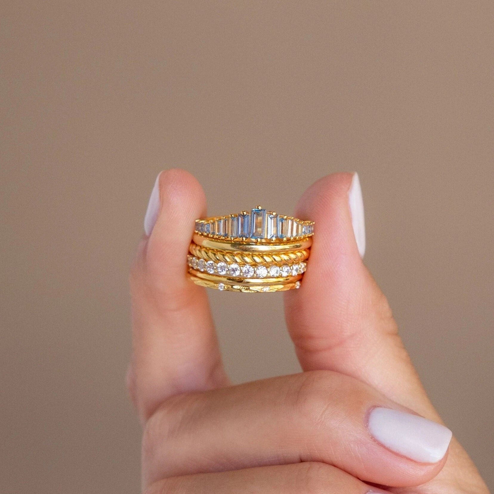 A hand with pale nails holds several stacked gold and diamond rings, including the Art Deco Aquamarine Ring, against a beige background for a hint of vintage glamour.