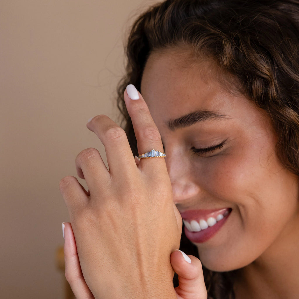 A smiling woman with curly hair shows off the Art Deco Aquamarine Ring, a vintage glamour gold ring featuring a princess-cut aquamarine and blue stones, on her hand near her face.