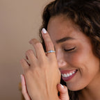A smiling woman with curly hair shows off the Art Deco Aquamarine Ring, a vintage glamour gold ring featuring a princess-cut aquamarine and blue stones, on her hand near her face.