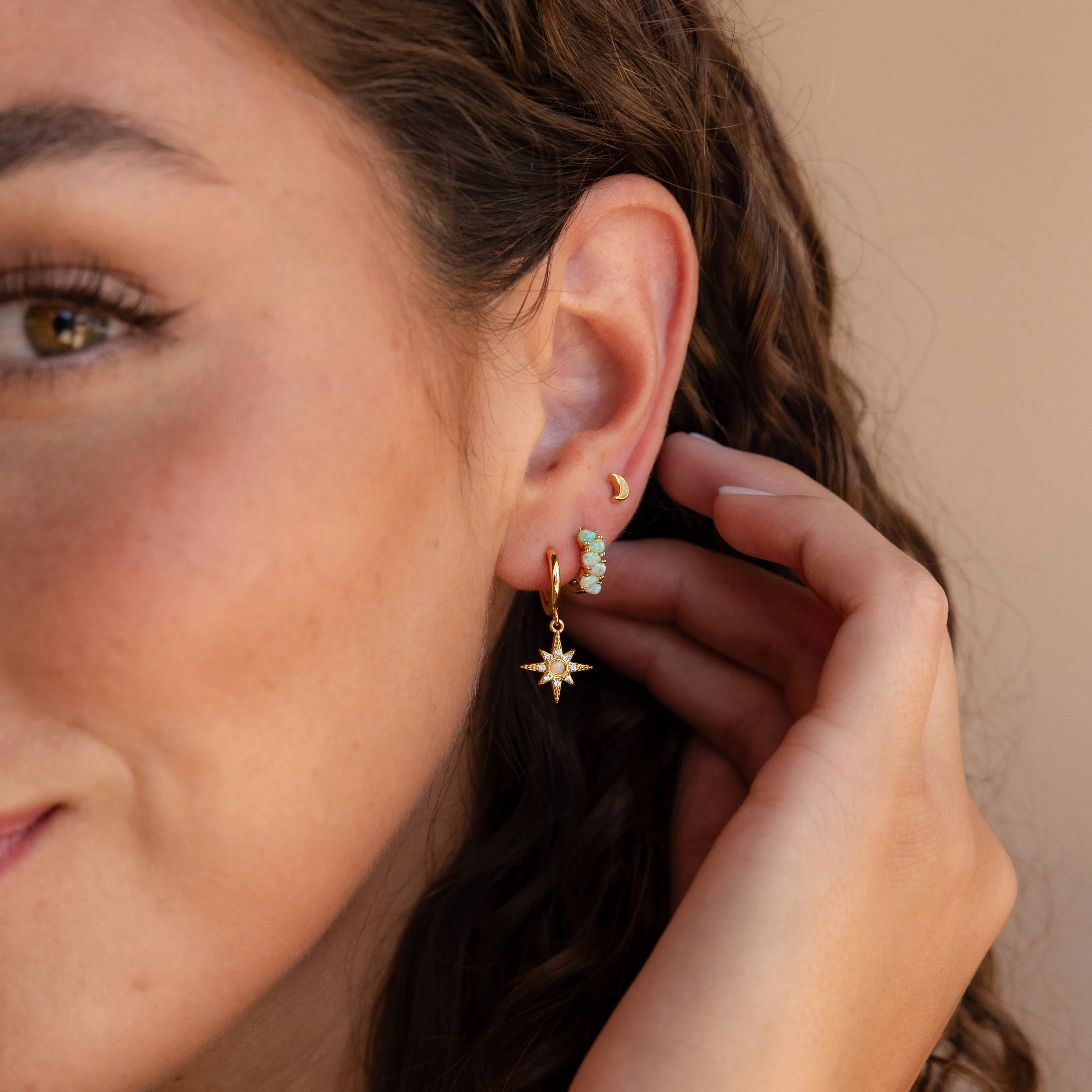 Woman with curly hair wearing four gold earrings, including Ombre Opal Huggies, a star-shaped hoop, iridescent teardrop opal earrings, and a small moon stud.