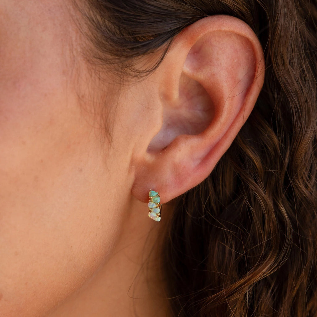 Close-up of a woman's ear with wavy hair, wearing Ombre Opal Huggies—small gold earrings adorned with iridescent opal stones.