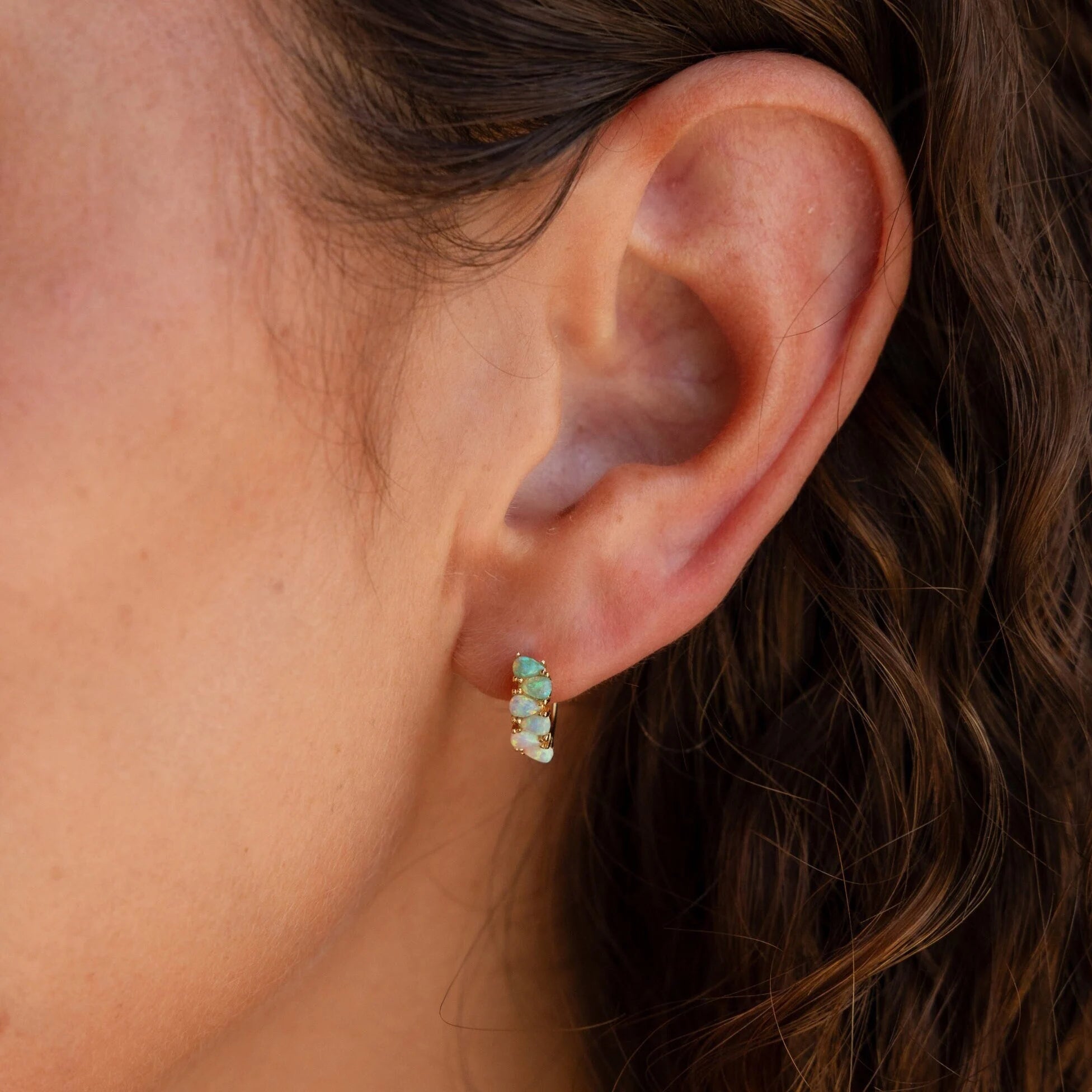 Close-up of a woman's ear with wavy hair, wearing Ombre Opal Huggies—small gold earrings adorned with iridescent opal stones.