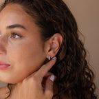 A woman with curly hair touches her ear, displaying Ombre Opal Huggies—delicate earrings that shimmer with iridescent hues in the light.
