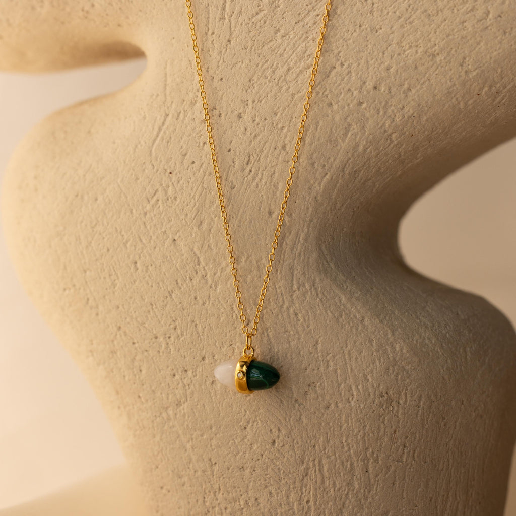 The Malachite & Moonstone Necklace features a green and white pendant, evoking healing gemstone jewelry, and is displayed on a textured abstract beige stand.