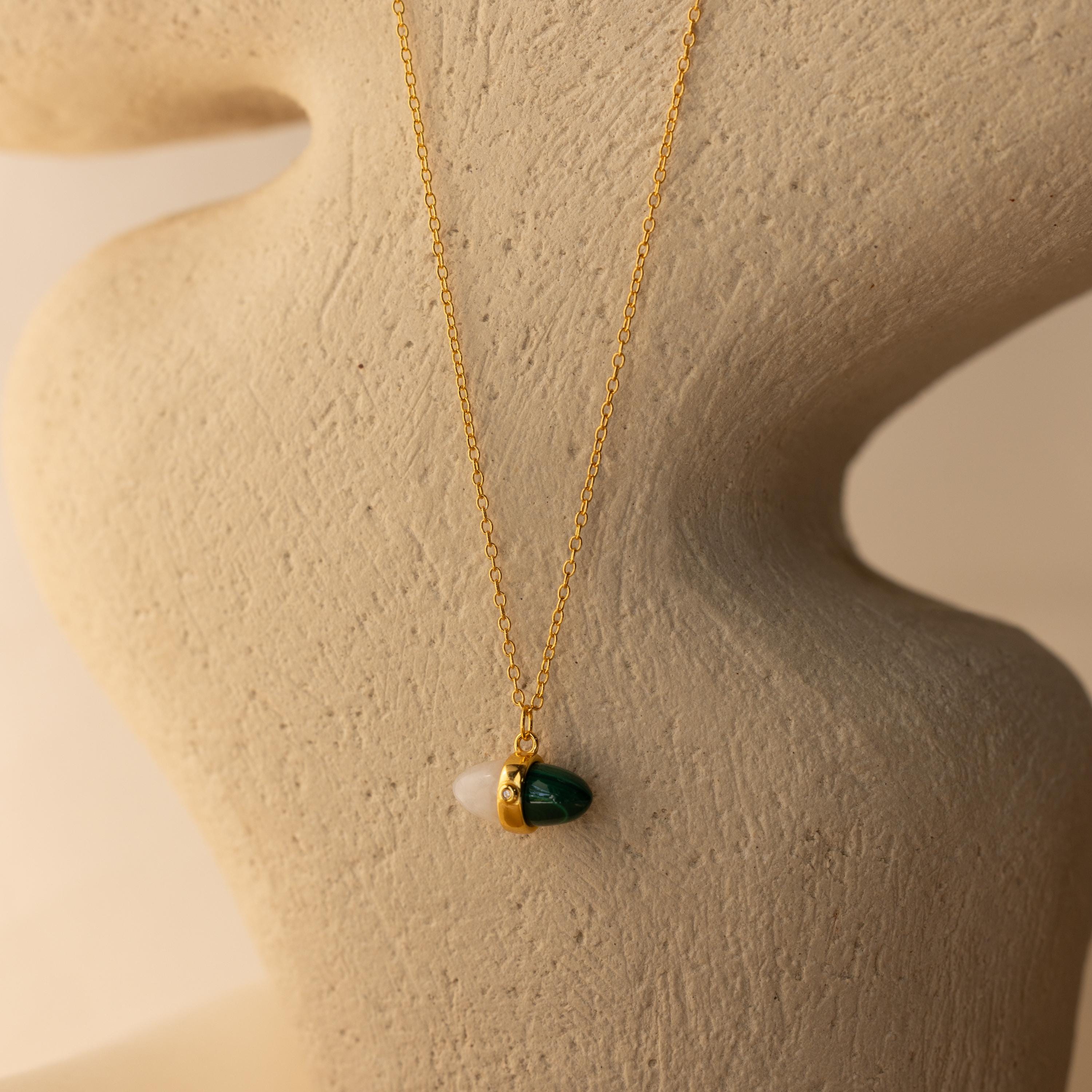 The Malachite & Moonstone Necklace features a green and white pendant, evoking healing gemstone jewelry, and is displayed on a textured abstract beige stand.