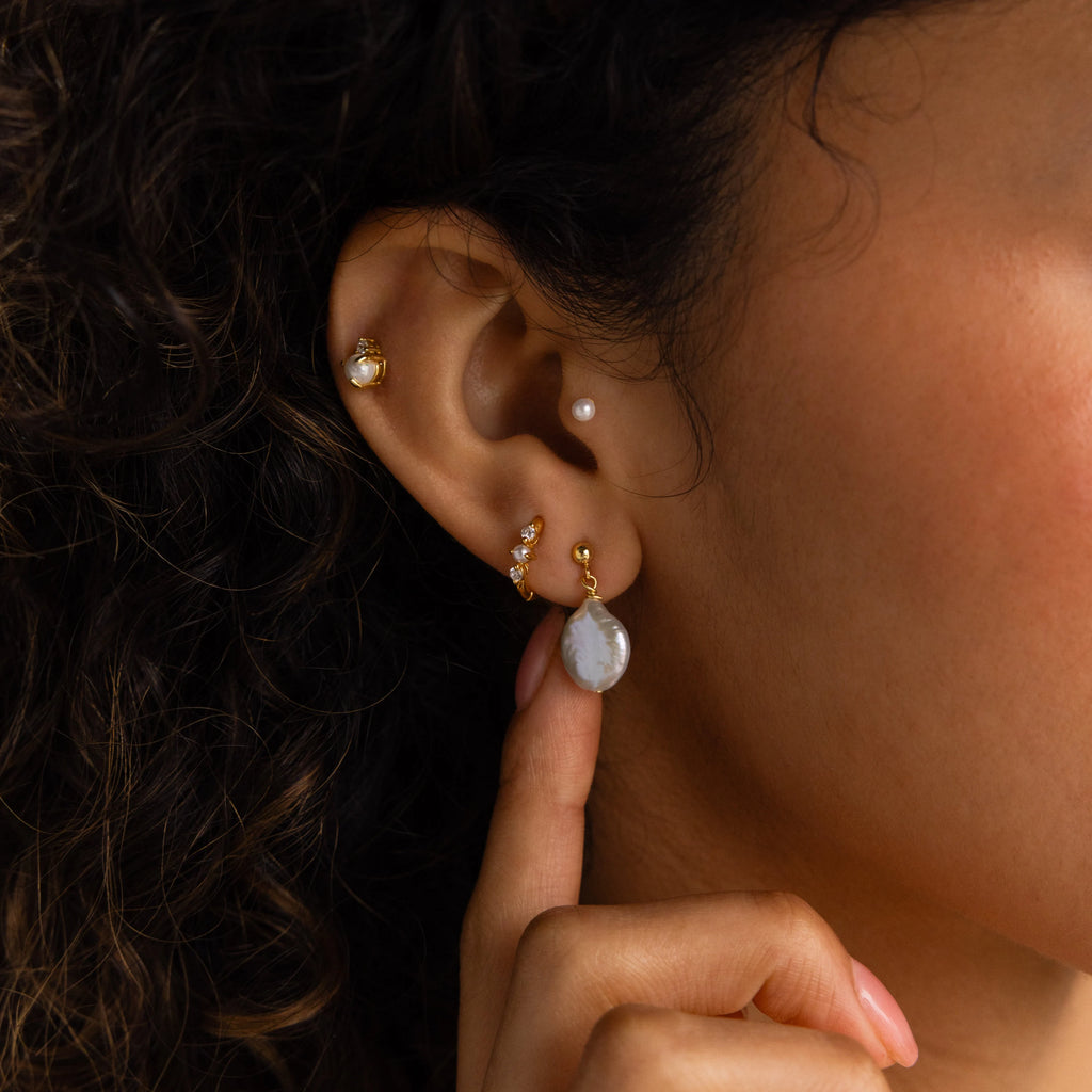 Close-up of a woman's ear wearing gold stud and Round Pearl Drop Earrings; she gently touches one earring with her finger.