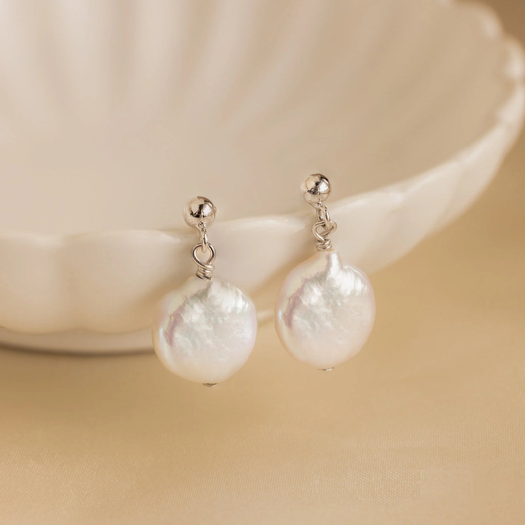 Round Pearl Drop Earrings in silver and white, displayed on a scalloped ceramic dish, add elegance to any outfit.