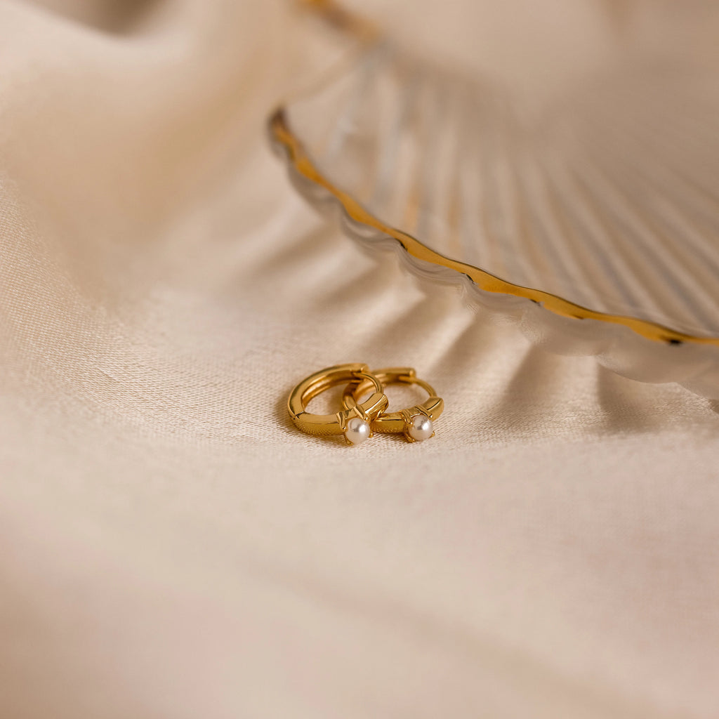The Tiny Pearl Huggies, two gold huggie hoops adorned with pearls, rest on soft beige fabric next to a decorative shell dish for an elegant display.