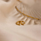 The Tiny Pearl Huggies, two gold huggie hoops adorned with pearls, rest on soft beige fabric next to a decorative shell dish for an elegant display.
