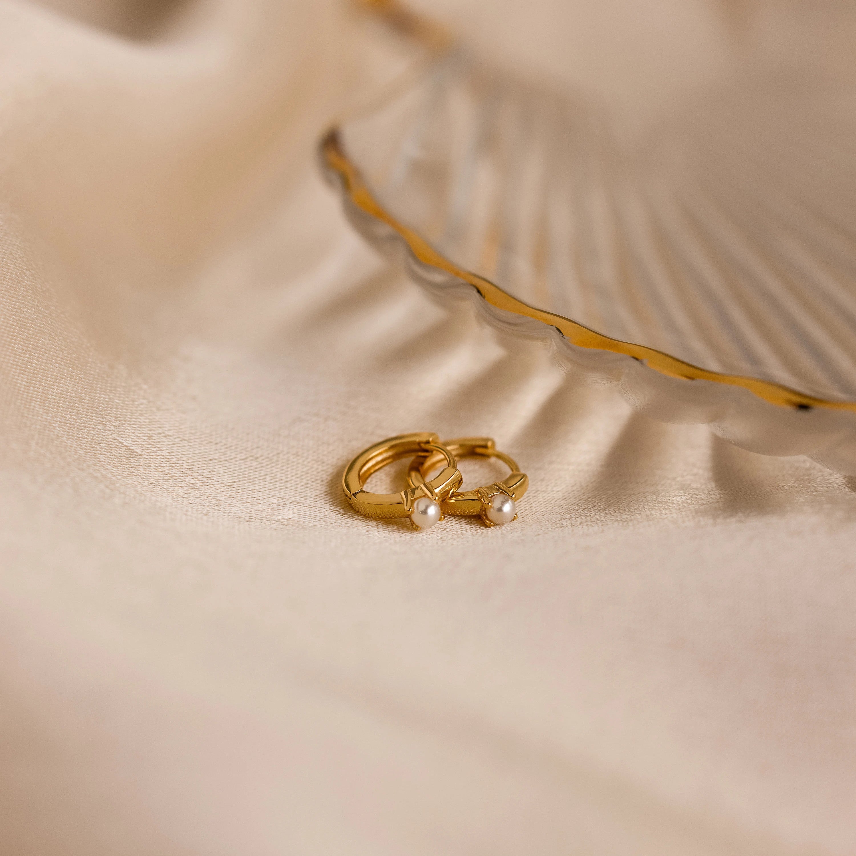 The Tiny Pearl Huggies, two gold huggie hoops adorned with pearls, rest on soft beige fabric next to a decorative shell dish for an elegant display.