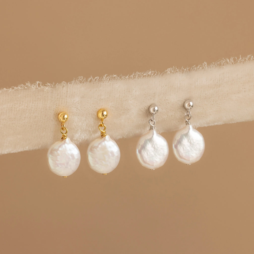 Two pairs of Round Pearl Drop Earrings—one with gold studs, the other with silver studs—are elegantly displayed on beige fabric.